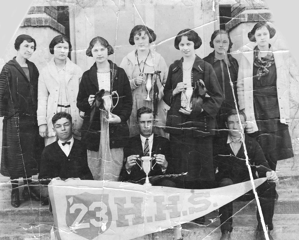 White students posing in front of school building with trophies and a banner.