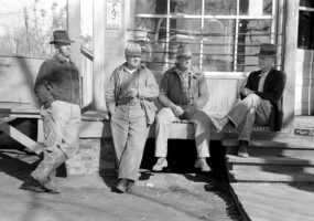 Four white men in hats lounging on porch of store