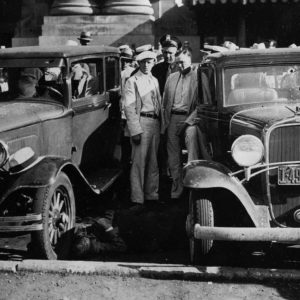 Large group of white men gathered around cars that have been shot