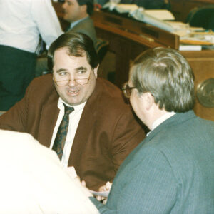 White man in glasses speaking with other white men in legislative chamber
