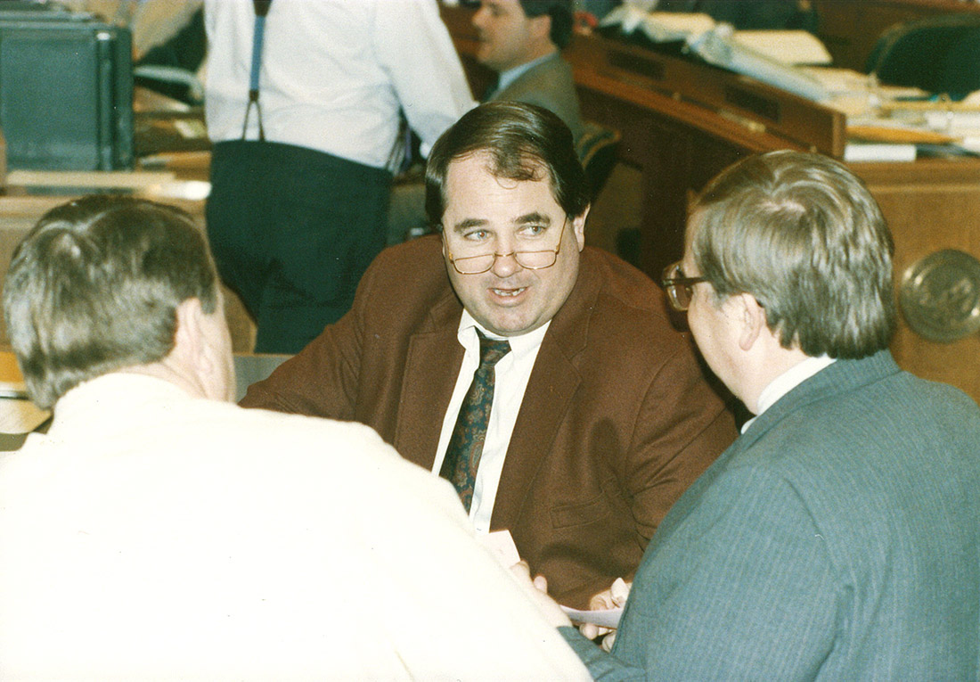 White man in glasses speaking with other white men in legislative chamber