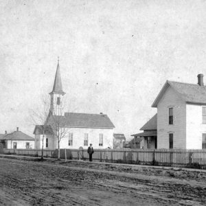 White wooden church buildings with dirt road and long fence in front with one man on the sidewalk