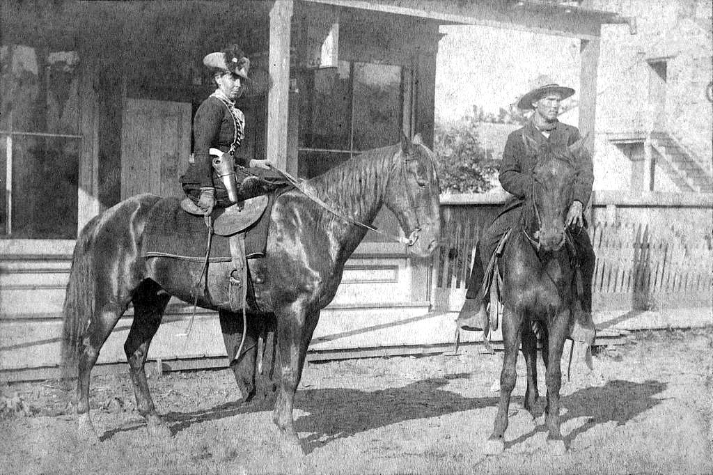White woman wearing a hat and a gun holster and white man each on horses