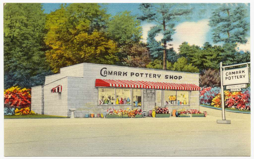 Postcard showing one story building with red and white striped awning and flowers in front and colorful pottery inside