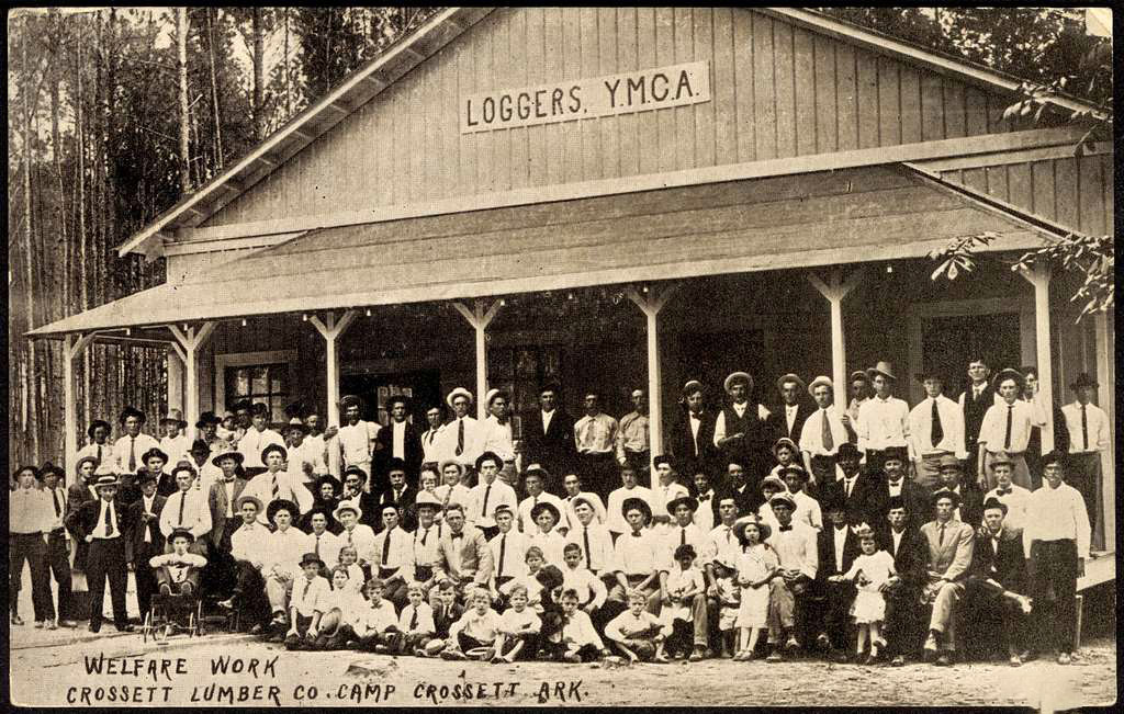 Large group of white adults and children in front of wooden building with sign saying "Loggers YMCA"