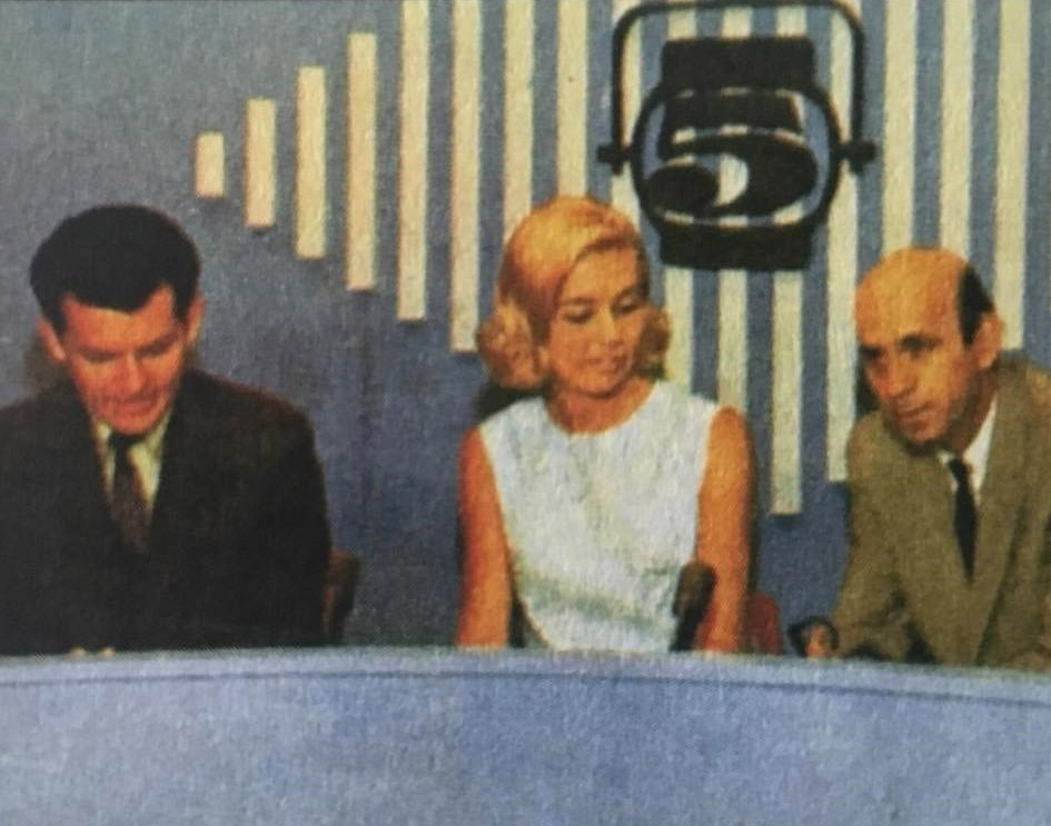 White woman in dress seated between two white men in television studio