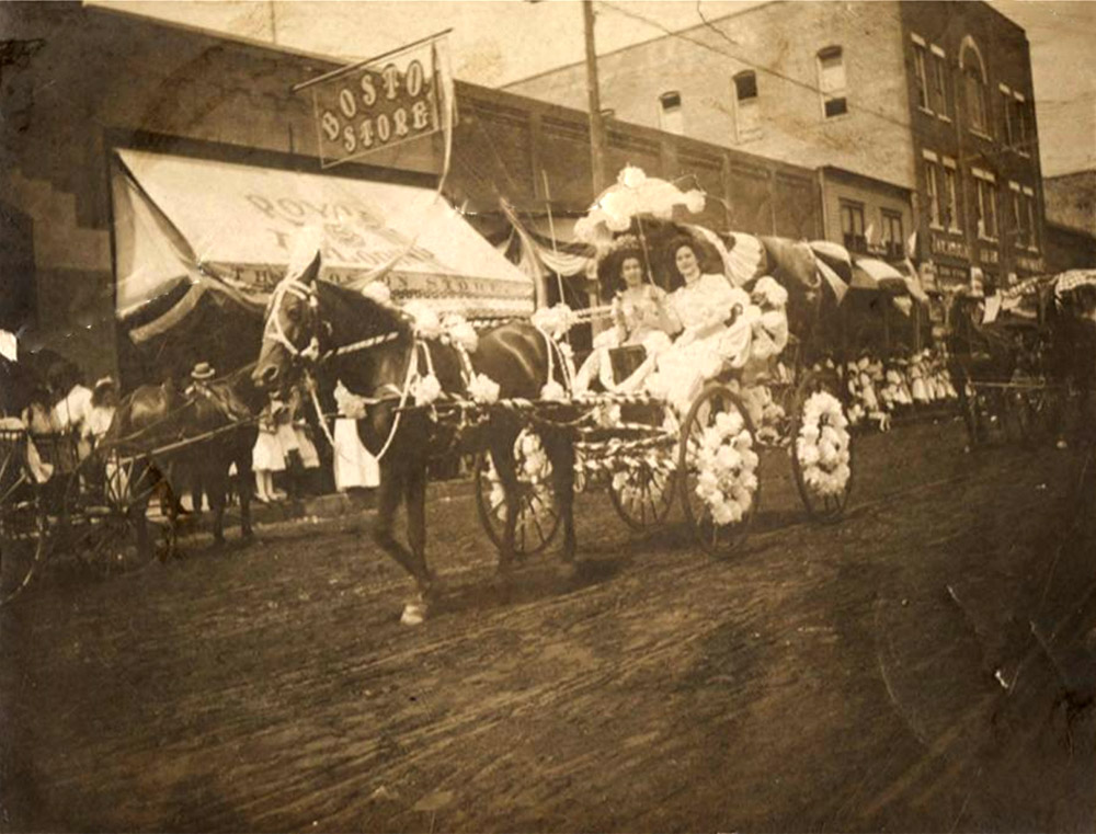 Two white women in decorated horse-drawn carriage proceeding down town street
