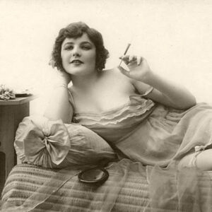 Scantily clad white woman reclining in bed smoking cigarette