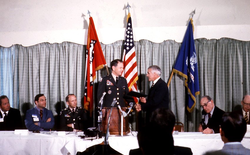 Two white men shaking hands at a lectern. Other white men seated at table