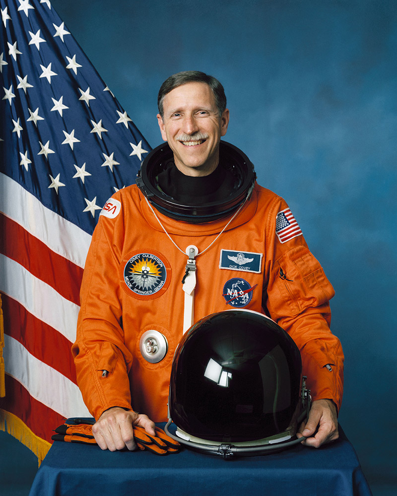 White man in astronaut uniform standing in front of American flag