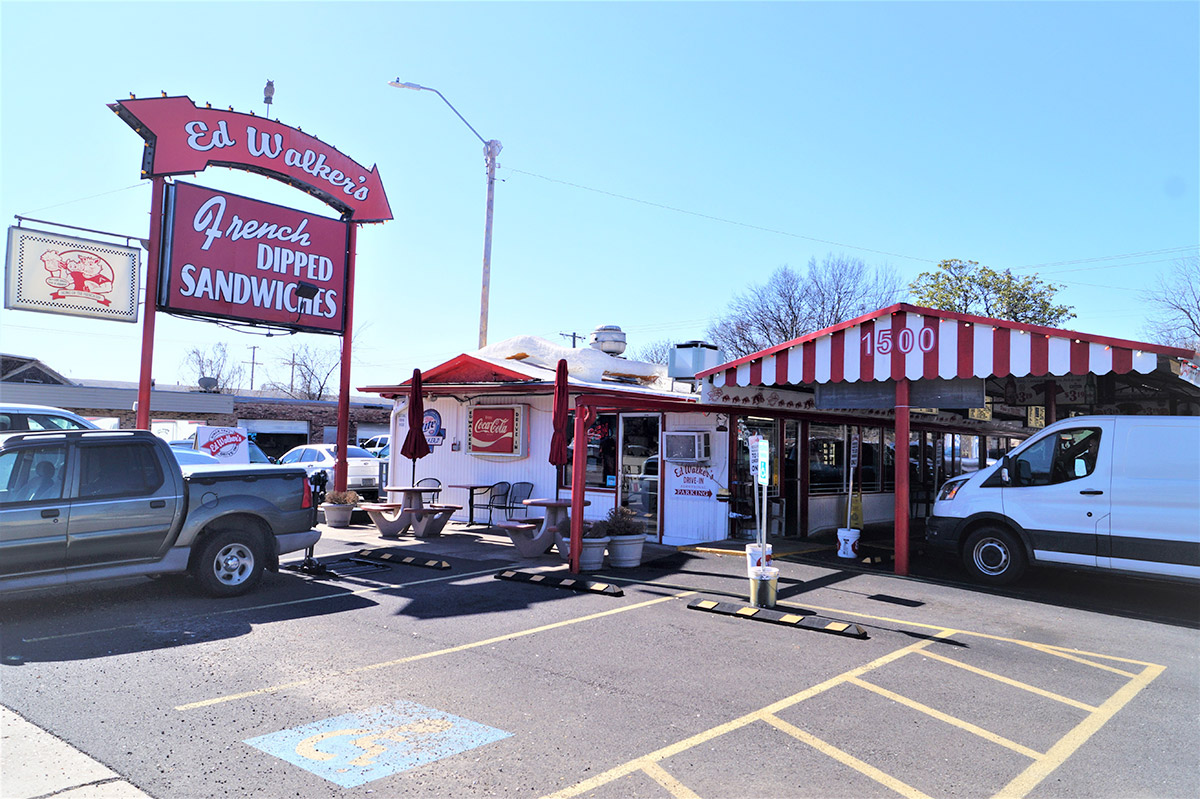 Cars parked beneath awning next to metal building with big red sign saying "Ed Walkers French dipped sandwiches"