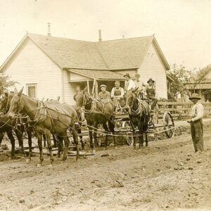 White men in wagon hitched to several horses with other white people standing about and houses in background