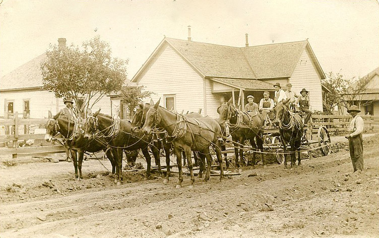 White men in wagon hitched to several horses with other white people standing about and houses in background