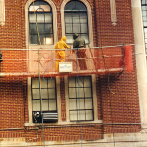 Two figures in protective suits standing on scaffold on multistory brick building