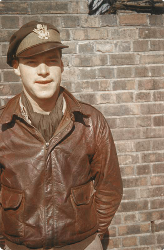 White man in military cap and leather jacket standing in front of brick wall