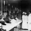 Line of white men with needles in their arms and tubes running up from them while white suited nurses observe