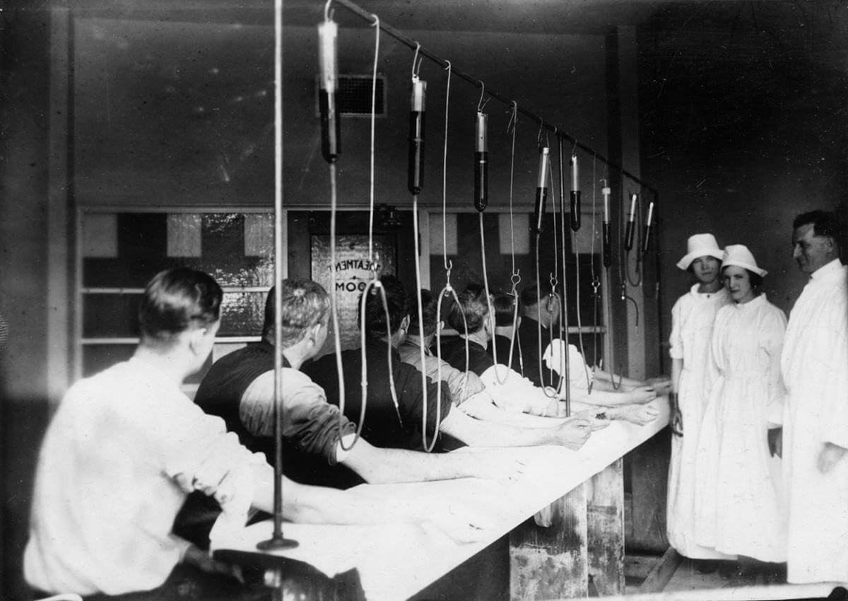 Line of white men with needles in their arms and tubes running up from them while white suited nurses observe