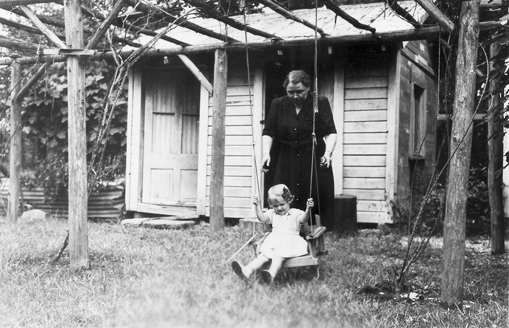 White woman in dress pushing white child in swing with small cottage in background