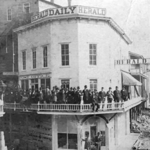 Large crowd of white men milling about by second floor exterior walkway of three-story building