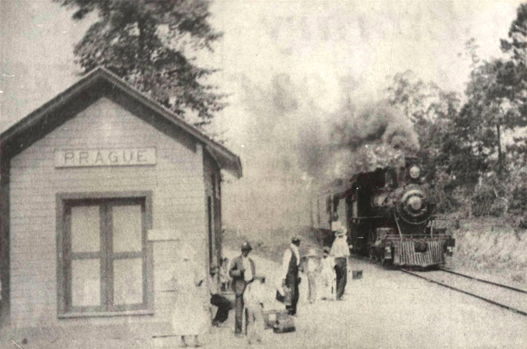 Small wooden building with sign saying "Prague" and several people waiting for arriving train