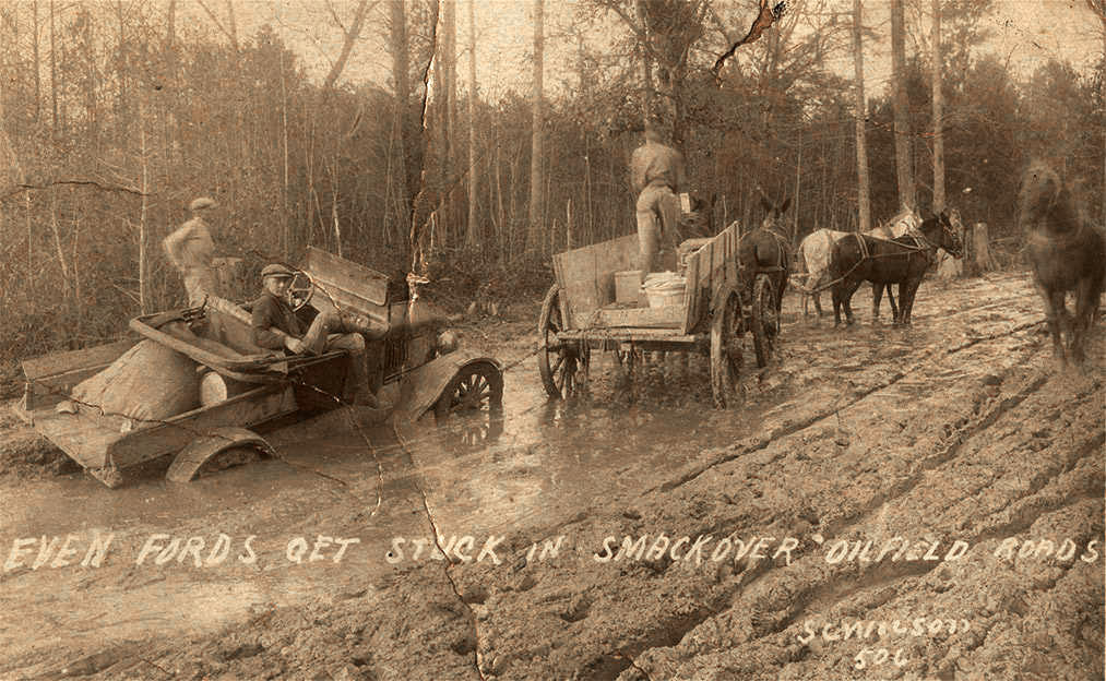 Car stuck in mud being towed by team of horses; writing on photo says "even fords get stuck in smackover oilfield roads"