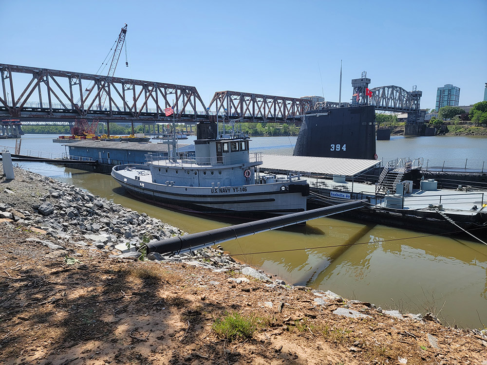 Tugboat and submarine docked on river with bridge in background