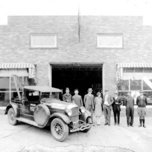 Group of white people standing next to car before brick building with central garage opening