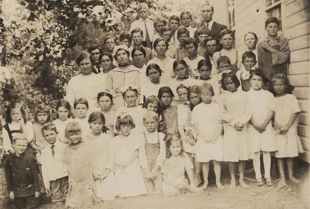 Large group of white children and adults beside wooden building