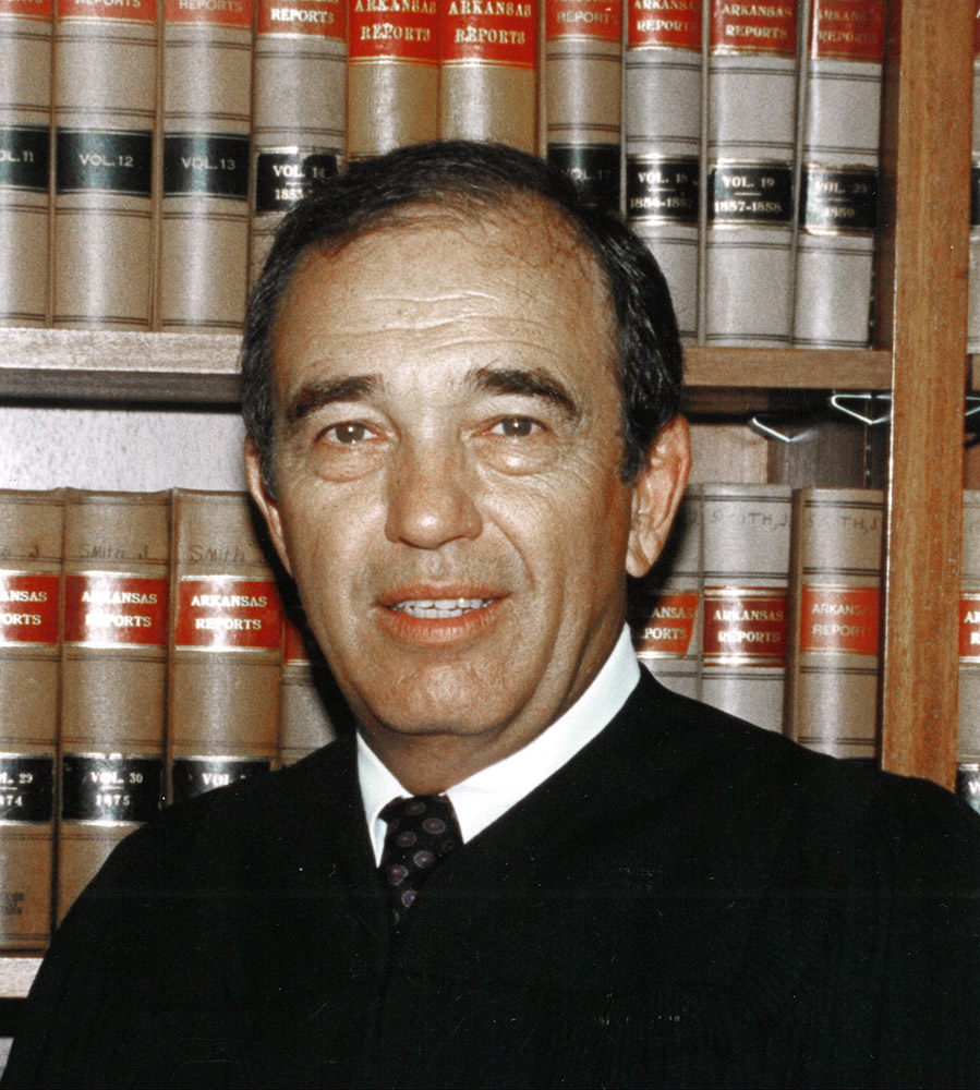 White man in tie and judge's robe standing in front of books
