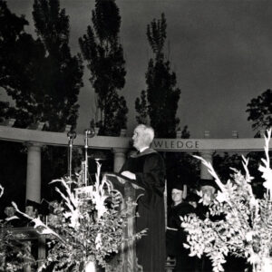 White man in academic robes on outdoor stage before microphone