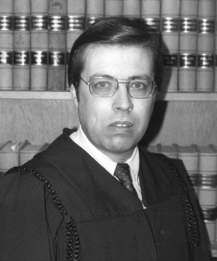 White man in glasses and judges robe standing in front of books