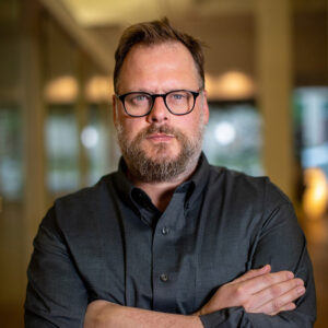 White man with beard and glasses wearing a dark button-down shirt