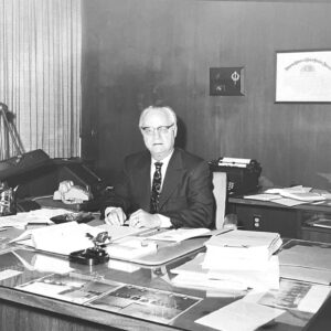 White man in suit and tie seated at cluttered desk