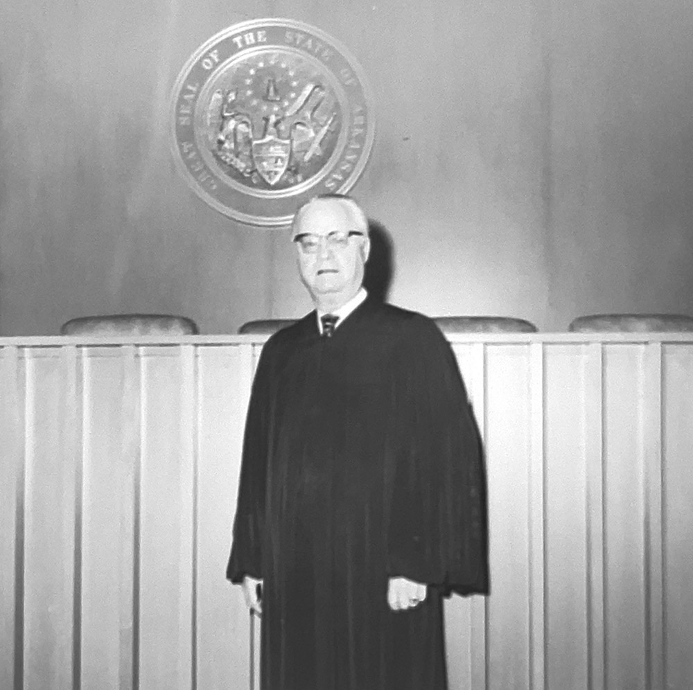 White man in judicial robes standing in front of judges' bench with state seal behind him