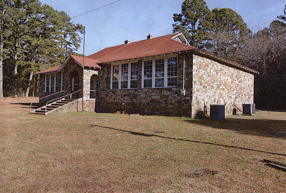 one-story rock school building with windows in front and red shingle roof and steps leading to front door