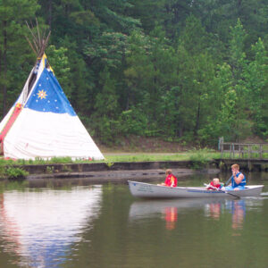 Group of three in a canoe paddling past a stylized tepee in a wooded area