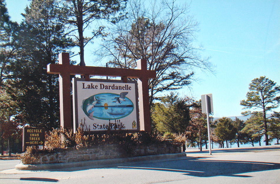 Large wooden sign reading "Lake Dardanelle State Park" next to trees and lake