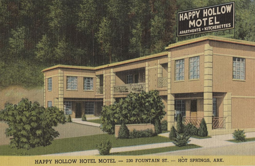 Two-story square building with multi-pane windows and sign on top saying "Happy Hollow Motel Apartments Kitchenettes"