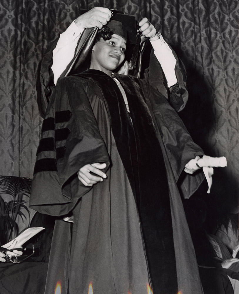 Young black woman in graduation robe while partially hidden white man puts academic hood around her neck