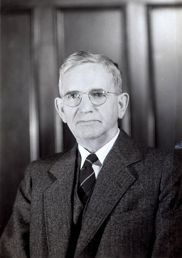 White man in suit and tie and glasses sitting at desk