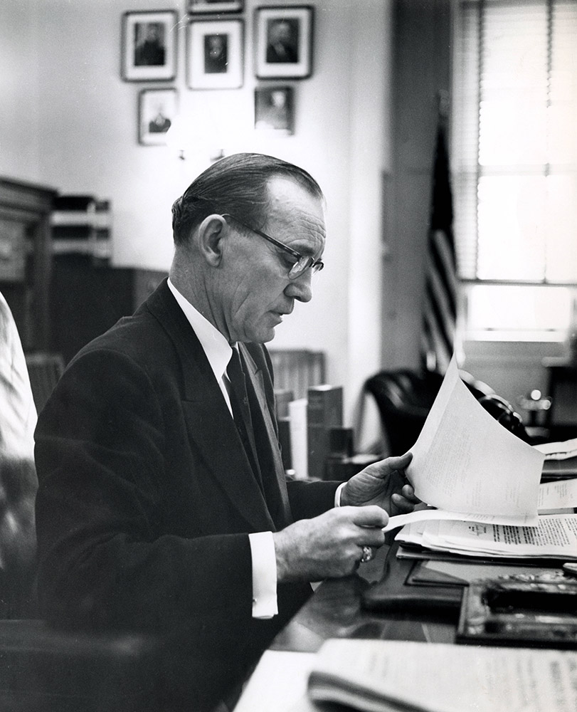 White man in suit and tie and glasses sitting at desk looking at papers