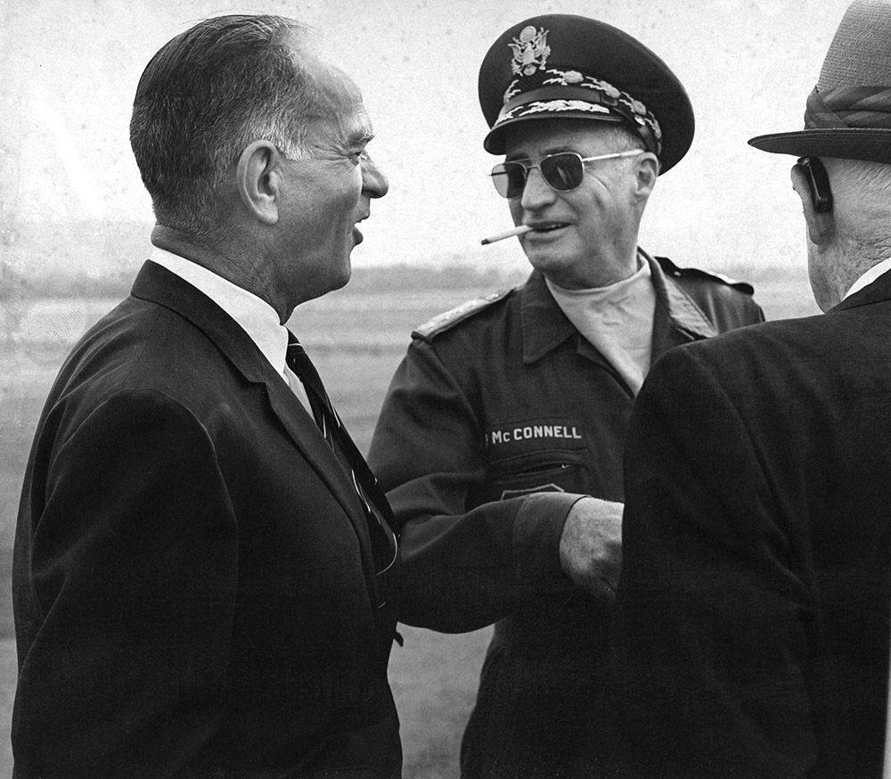 White man in military garb and a cigarette sticking out of his mouth standing between two white men in suits