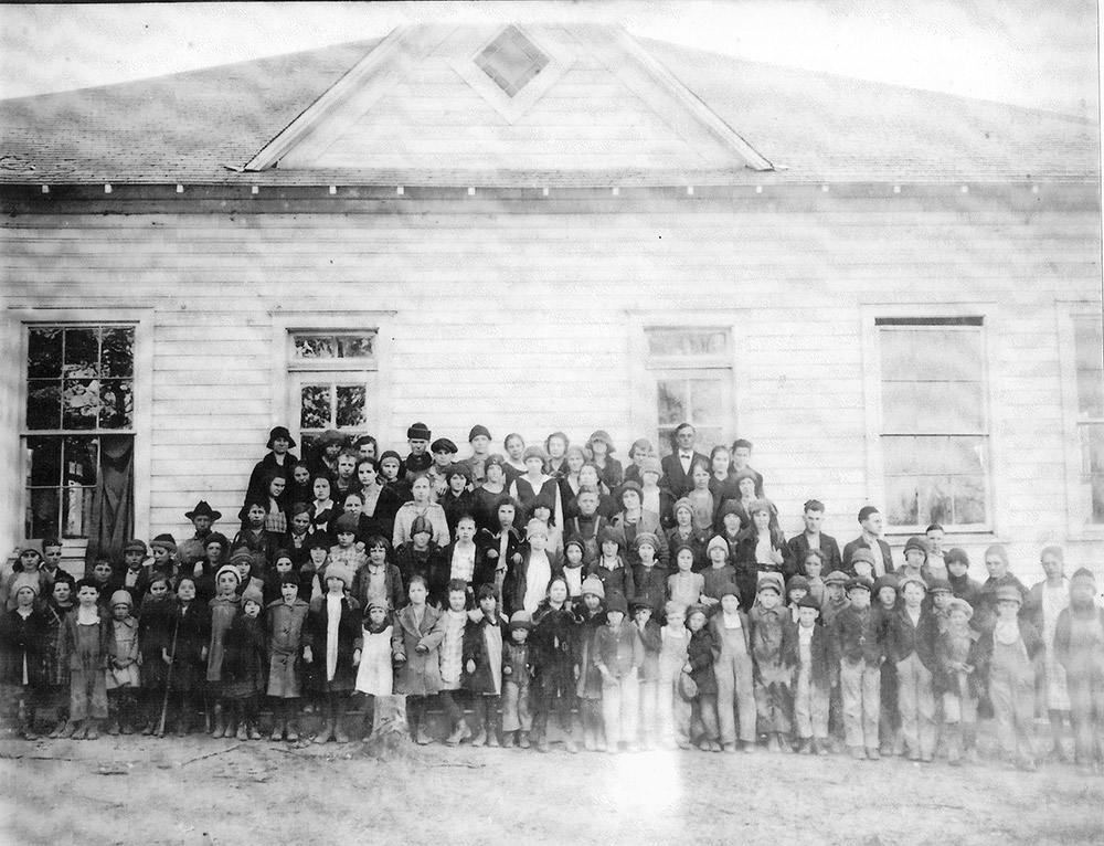 Large group of people standing before single story white wooden building