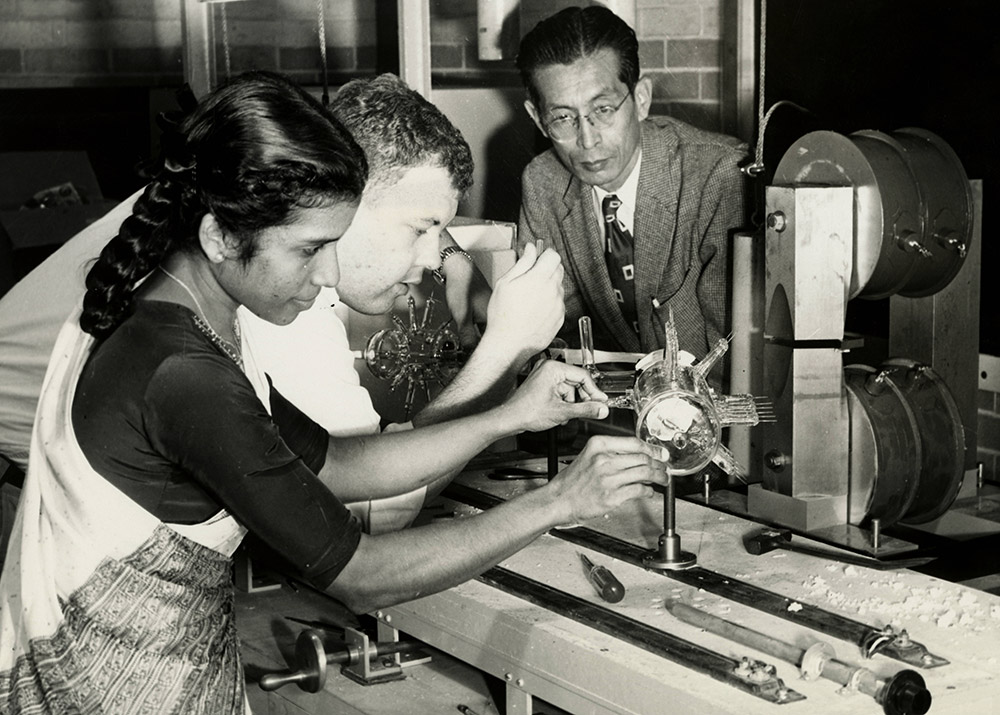 Asian man in suit and tie overseeing a male and a female student as they operate a piece of laboratory equipment