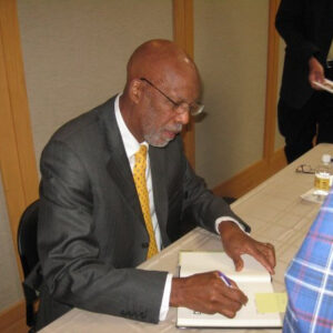 African American man in suit and tie seated at table signing a book