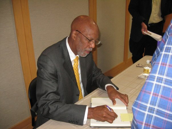 African American man in suit and tie seated at table signing a book
