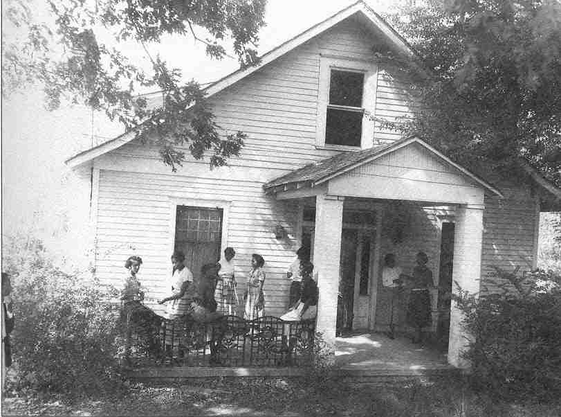 Multistory white wooden house with African Americans sitting and standing on the porch