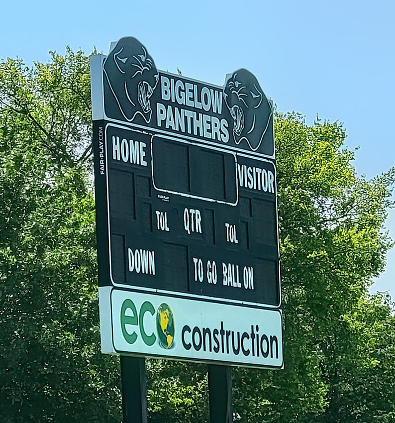 Metal scoreboard saying "Bigelow Panthers" on top with trees in background