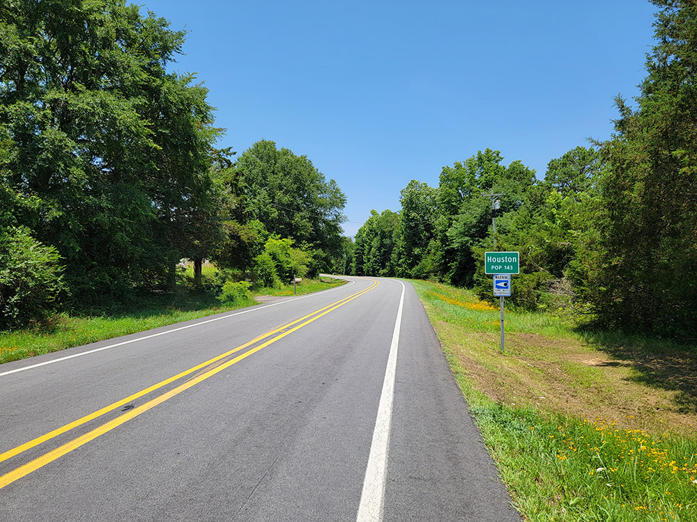 Rural highway with trees on either side and green sign saying "Houston population one hundred forty-three"