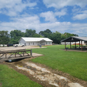 Single story white wooden building adjacent to park area with picnic tables and a wooden bridge across a creek